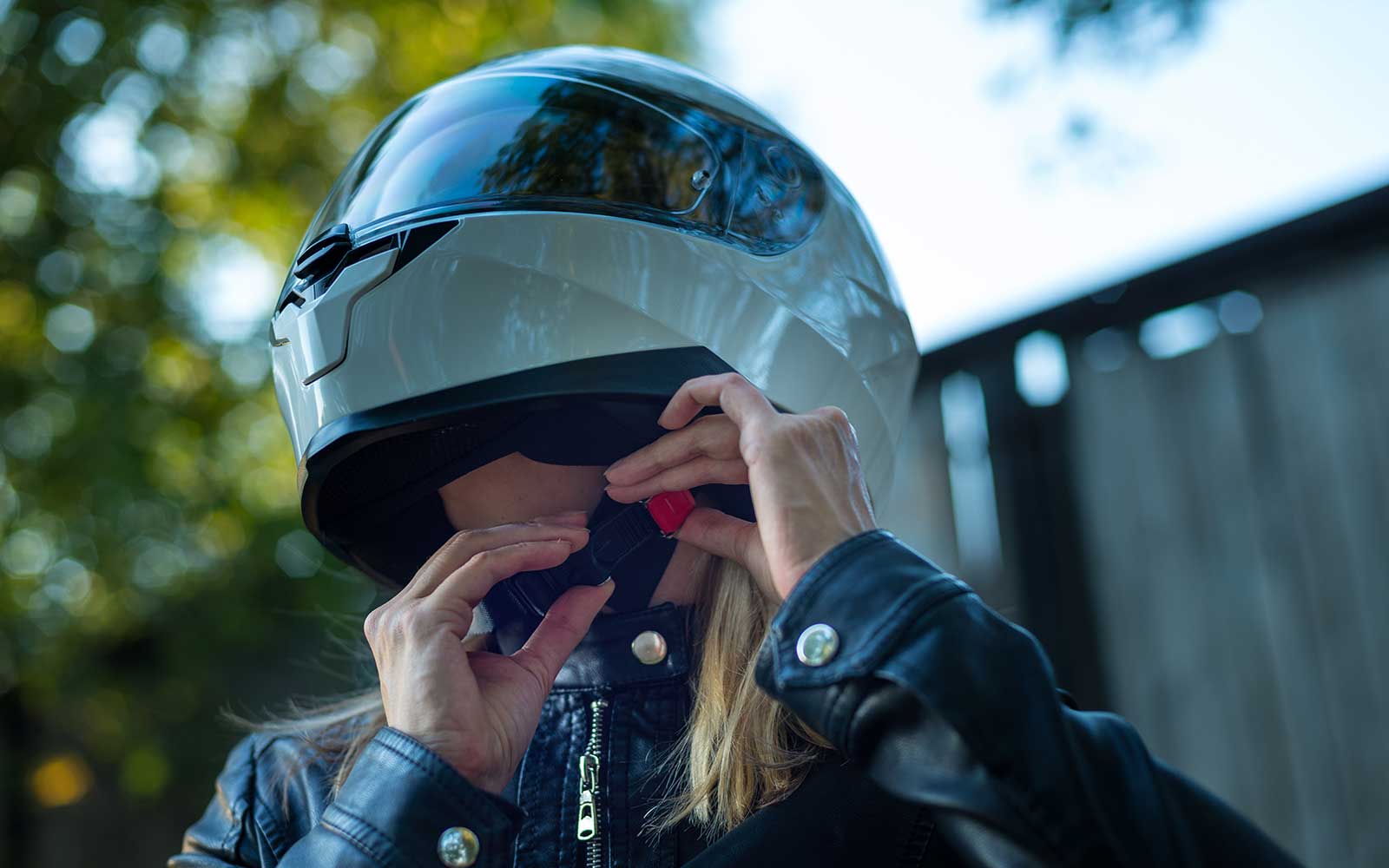 Eine Motorradfahrerin in einer schwarzen Lederjacke prüft den Sitz ihres weißen Integralhelms.