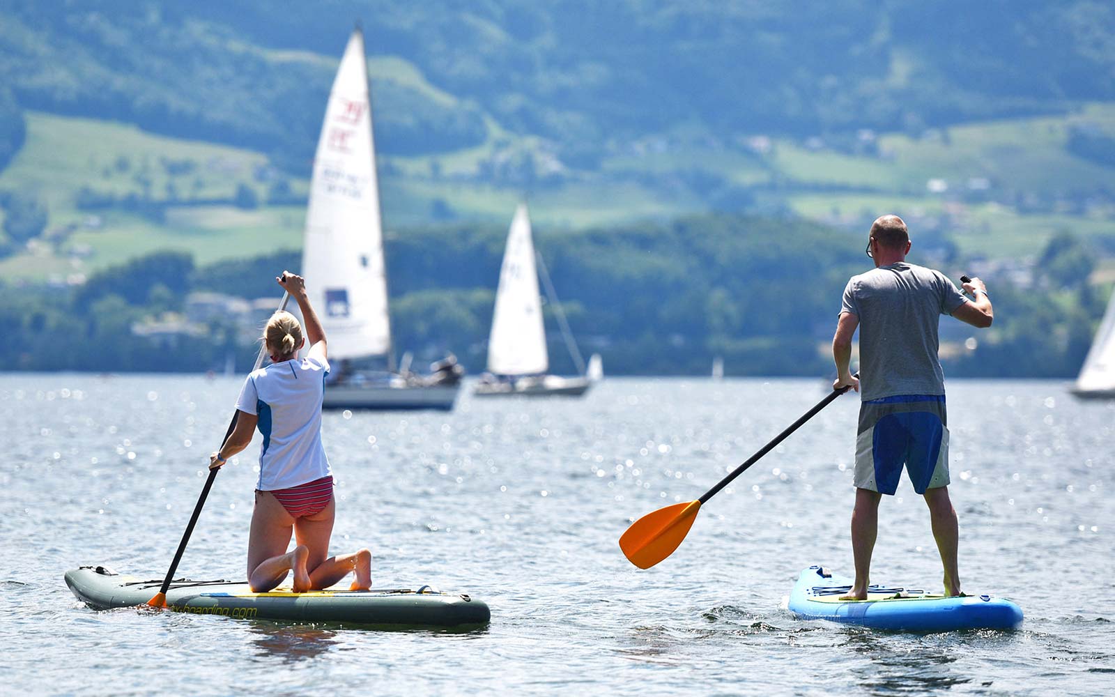 Zwei Stand Up Paddler:innen sind am See unterwegs. Vor ihnen sind mehrere Segelboote.